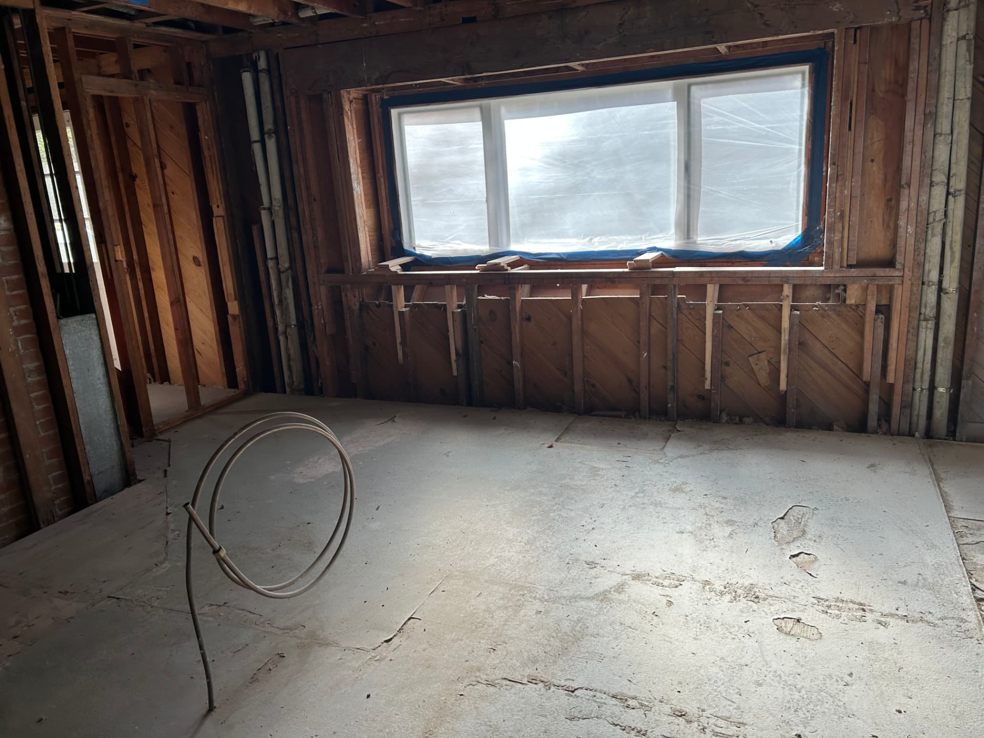 During kitchen rebuild: walls opened to exposed studs and bare subfloor, window masked with plastic and tape