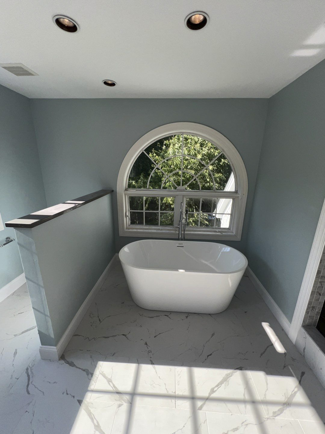 After bathroom work: freestanding white bathtub beneath an arched window, blue walls and marble-look floor tile